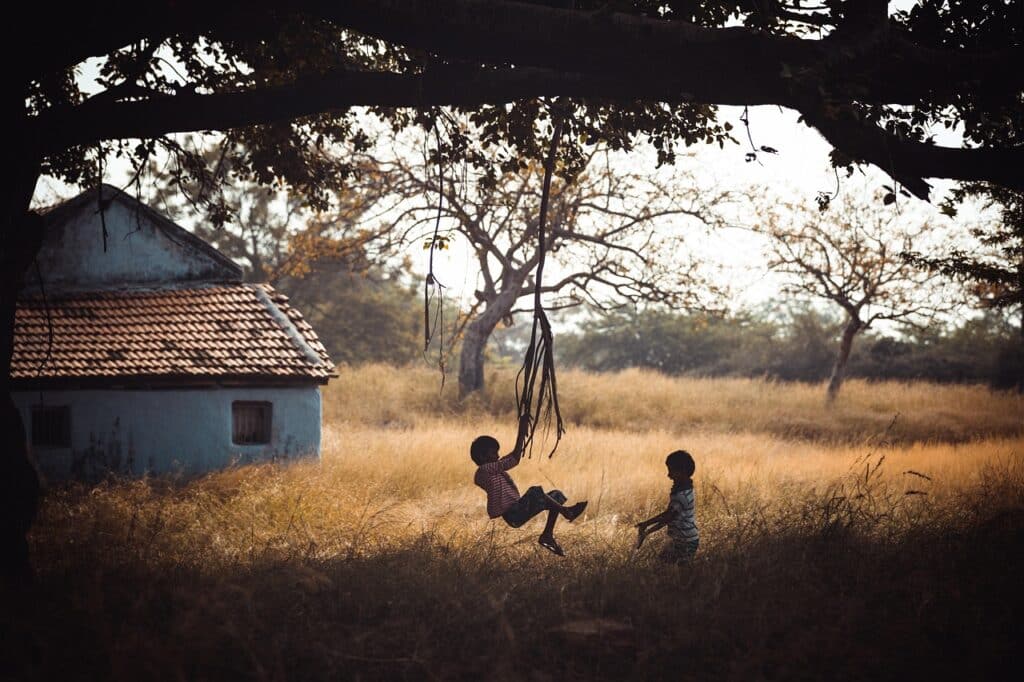 Two young children play under a large tree near a small house, one swinging from a rope in a sunny field.